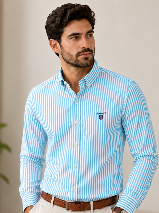 A man wearing a light blue and white vertically striped long-sleeve button-down shirt by Glandex, paired with beige trousers and a brown belt, standing in a brightly lit indoor setting.